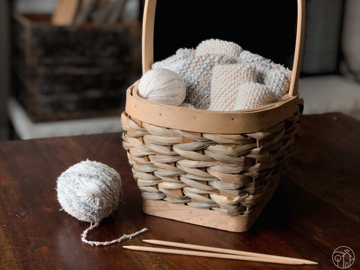 A light wood basket sits on a dark wood table. The basket is full of rolled knitted cloths, with a ball of white yarn to the front left. On the table is another neutral-coloured ball of cotton yarn and a pair of birch wood double-pointed knitting needles.